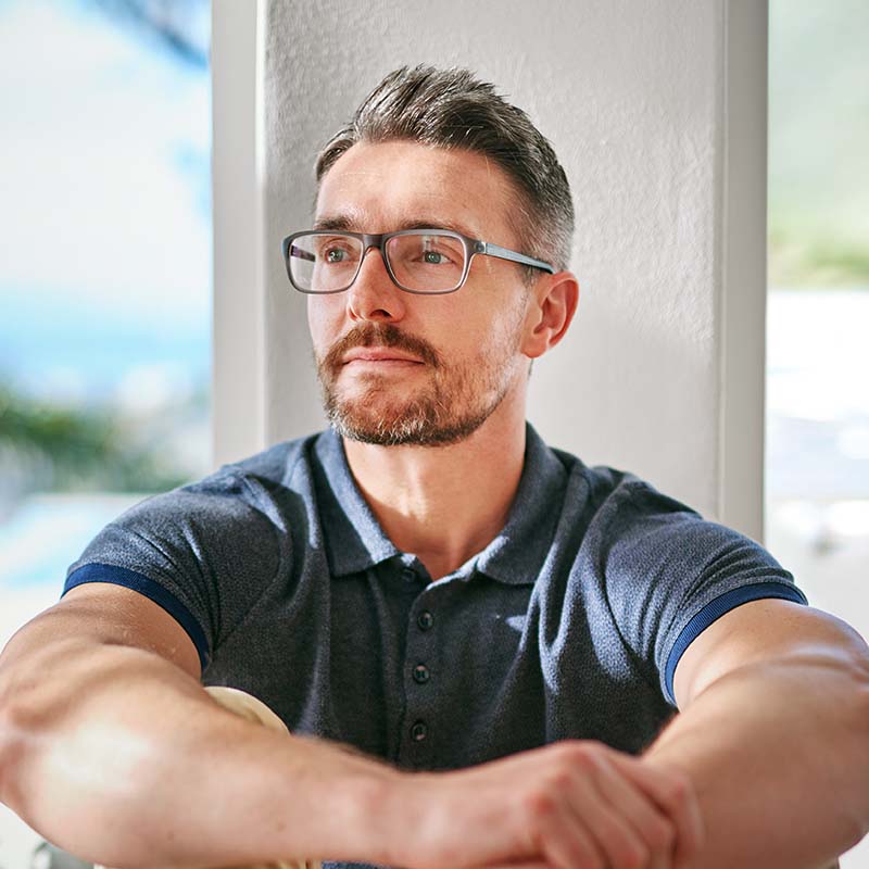 Mann mit Brille in blauem Poloshirt sitzt nachdenklich an einem Fenster mit Ausblick auf Landschaft